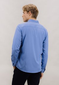 Lyseblå skjorte med lange ermer laget av glatt stoff. Har krage med knapper og mansjetter. Inkluderer sømdetaljer og avrundet nederkant.