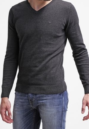 Mężczyzna w ciemnoszarym swetrze z dekoltem w serek i niebieskich dżinsach, stojący z lekko wyciągniętymi ramionami na białym tle.