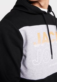 Felpa nera e grigia con tasca a marsupio. Presenta un logo arancione e bianco, e un cappuccio con cordini. Realizzata in morbido cotone.