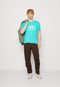 Türkises T-Shirt mit weißem Logo, braune Cargohosen mit seitlichen Reißverschlüssen, hält eine grüne Steppjacke, beiges Schuhwerk.