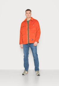 Νεαρός άνδρας φορώντας πορτοκαλί μπουφάν, πράσινο μπλουζάκι, μπλε τζιν και μπεζ αθλητικά παπούτσια, που στέκεται μπροστά σε απλό ανοιχτό γκρι φόντο.