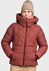 Rote gesteppte Daunenjacke mit hochgezogenem Kragen, Frontreißverschluss und zwei Seitentaschen. Getragen mit einer beigen Strickmütze.