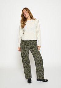 Krémový textúrovaný sveter s dlhými rukávmi; zelené široké nohavice s leopardím vzorom; čierne topánky, stojace na čistom bielom pozadí.