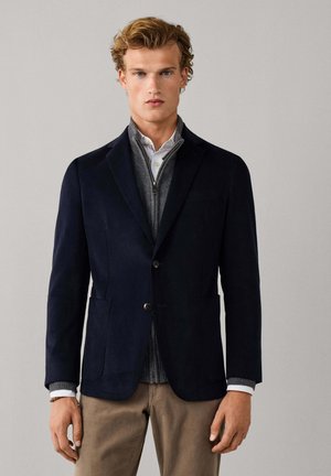 Junger Mann mit blondem lockigem Haar, der einen marineblauen Blazer, einen grauen Reißverschluss-Pullover, ein weißes Hemd und beige Hosen trägt, steht vor einem einfarbigen Hintergrund.