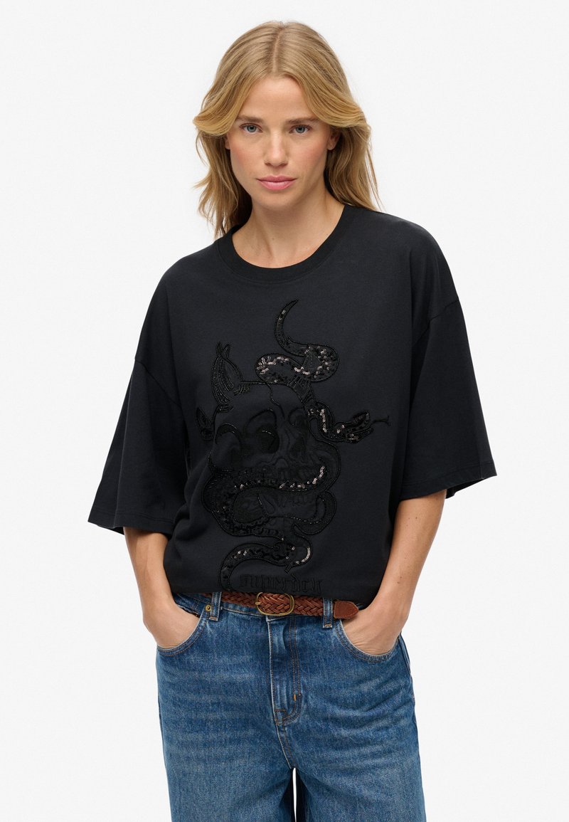 Camiseta negra de gran tamaño con un diseño bordado texturizado oscuro que presenta un patrón de calavera y serpiente. Combinada con jeans azules y un cinturón marrón.
