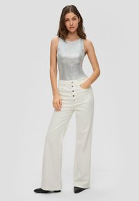 Zilveren geribbelde tanktop met witte hoge taille wijde pijpenbroeken met vijf knopen. Zwarte enkellaarsjes. Eenvoudige achtergrond.