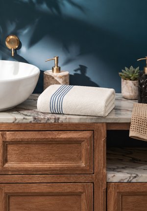 Serviette blanche pliée avec des rayures bleues sur un plan de travail en marbre avec un évier blanc, un distributeur de savon doré, une petite plante et une coiffeuse en bois en dessous.