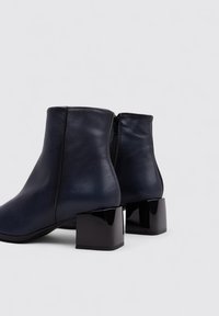 Botas de tobillo de cuero azul marino con una textura suave, que cuentan con cierre de cremallera y tacones cuadrados brillantes. Diseño simple con líneas limpias.