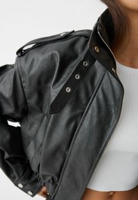 Veste en cuir synthétique noire avec un col montant, fermeture éclair argentée et boutons-pression. Présente un design court avec des détails de couture élégants.
