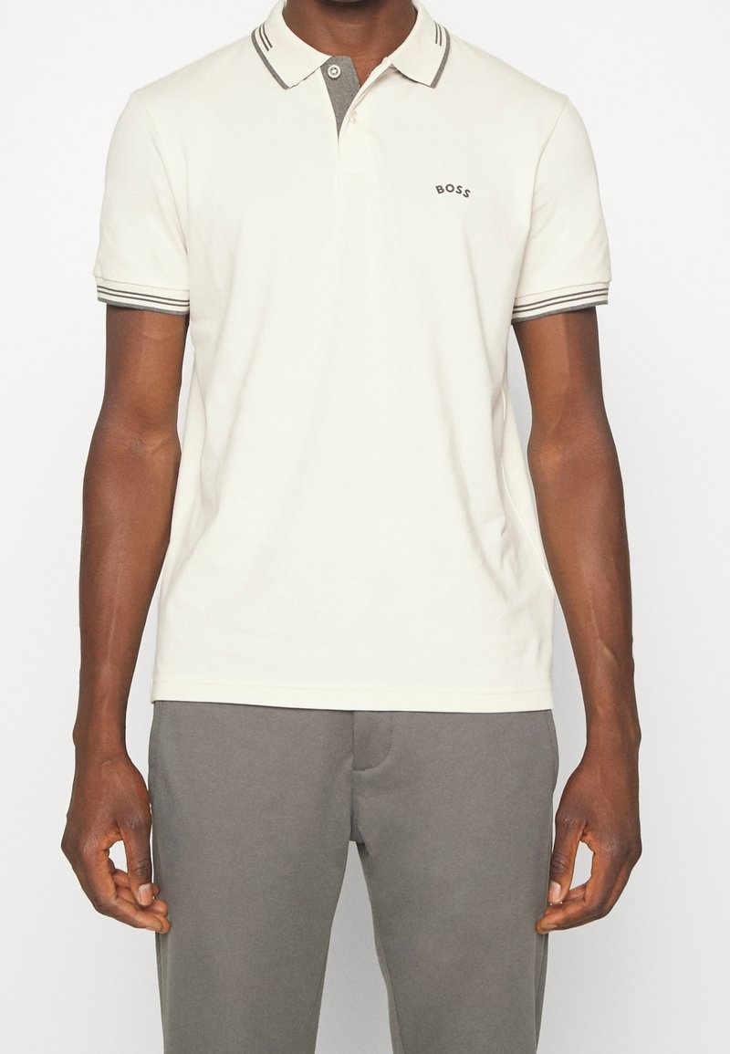 Homme portant un polo blanc à manches courtes avec col et poignets à double rayure, logo "BOSS" sur la poitrine, associé à un pantalon gris.