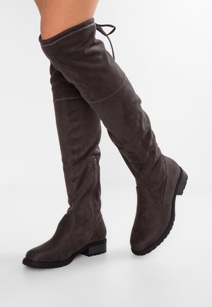 Mujer que lleva botas de ante gris oscuro hasta la rodilla con cremallera lateral y tacón bajo, de pie contra un fondo blanco.