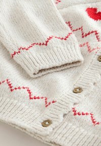 Knitted cardigan in cream with red zigzag and heart patterns. Features ribbed cuffs and brown buttons. Soft, textured material.