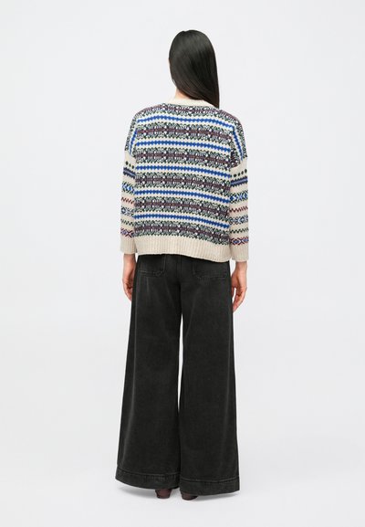 Person with long dark hair wears a patterned knit sweater and wide-leg black pants, standing facing away against a plain background.