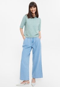 Maglione lavorato a maglia verde chiaro con maniche corte e dettagli in perline, abbinato a jeans a vita alta blu chiaro con gamba ampia e scarpe bianche a punta.