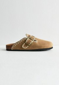 Brown suede slip-on shoe with a leather sole, featuring a decorative stitch pattern and a metallic buckle accent.