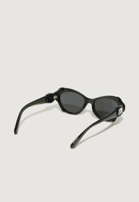 Gafas de sol negras con forma curvada de ojo de gato. Presentan lentes oscuros y montura de plástico brillante, acentuada por un detalle de logo metálico.