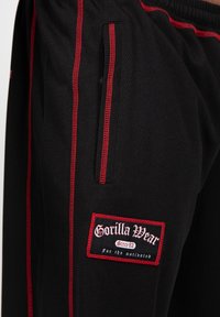 Zwarte sportbroek met rode zijstrepen en een label van stof met het "Gorilla Wear"-logo. Het materiaal lijkt textuur te hebben en ademend te zijn.