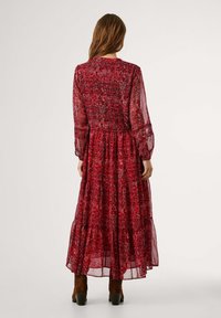 Lång, mjuk klänning i en röd blommig utskrift. Har genomskinliga ärmar, en figursydd bodice och en lager-på-lager volangslut. Bärs med bruna ankelstövlar.