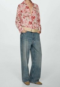 Blusa a fantasia floreale nei toni del rosa, maniche lunghe, abbinata a jeans a gamba larga di colore blu chiaro e una cintura beige. Scarpe lucide color cuoio a punta.