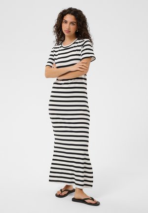 Mujer con cabello rizado que lleva un vestido largo a rayas horizontales en blanco y negro y chanclas negras, de pie con los brazos cruzados.