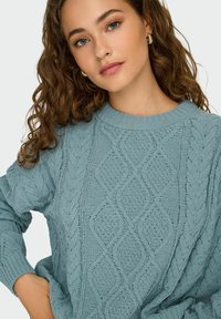 Pull en tricot bleu clair avec un motif en diamant et des détails en torsades. Encolure et poignets côtelés. Texture douce, coupe décontractée.