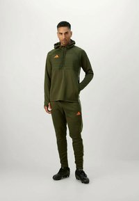 Sudadera deportiva verde oliva con cuello con cremallera y acentos naranjas, complementada con joggers a juego; ambos hechos de una tela suave y elástica.