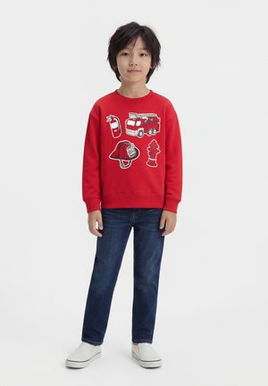 Garçon aux cheveux noirs courts portant un sweat-shirt rouge avec des motifs de camion de pompiers, d'extincteur, de casque et de borne d'incendie, un jean bleu et des chaussures blanches sans lacets.