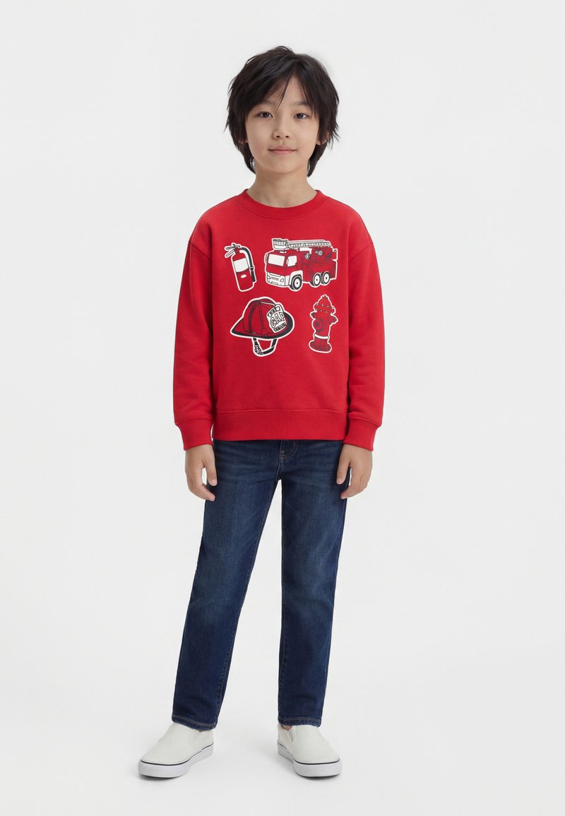 Garçon aux cheveux noirs courts portant un sweat-shirt rouge avec des motifs de camion de pompiers, d'extincteur, de casque et de borne d'incendie, un jean bleu et des chaussures blanches sans lacets.