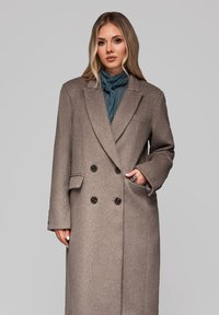 Brun uldblanding dobbeltbrystet frakke med en nakkekrage, to forreste lommer og store sorte knapper. Matcher med et teal-farvet tørklæde.