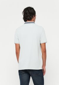 Mann ikledd i lys grå kortermet polo med marineblå striper på kragen, sammen med mørkeblå jeans, stående mot hvit bakgrunn.