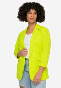 Blazer jaune fluo avec une coupe ajustée, revers à cran et deux poches avant, associé à un haut blanc et un short en denim déchiré.