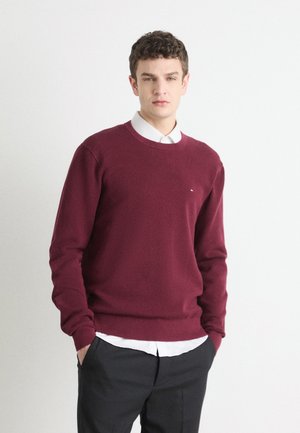 Maglione in maglia borgogna con scollatura rotonda, tessuto testurizzato e polsini a costine, indossato sopra una camicia bianca con colletto. Modello con pantaloni neri.