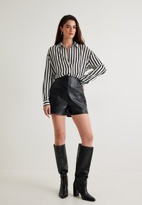 Pantalones cortos de cuero negro de cintura alta combinados con una camisa de botones a rayas en blanco y negro; modelo usando botas negras hasta la rodilla.