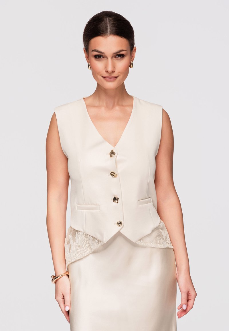 Femme aux cheveux foncés coiffés en arrière portant un gilet crème sans manches avec des boutons dorés et une jupe assortie, posant devant un fond uni.