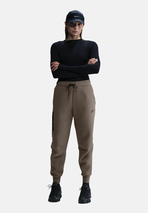 Maglietta nera a maniche lunghe, pantaloni jogger marroni con elastico alle caviglie e cordino. Accenti neri sui pantaloni jogger. Un cappellino nero e delle scarpe sportive nere completano il look.