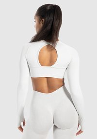 Vrouw met een hoge paardenstaart, gekleed in een witte lange mouw crop top met een uitsnede op de rug en bijpassende hoge taille leggings, staande tegen een grijze achtergrond.