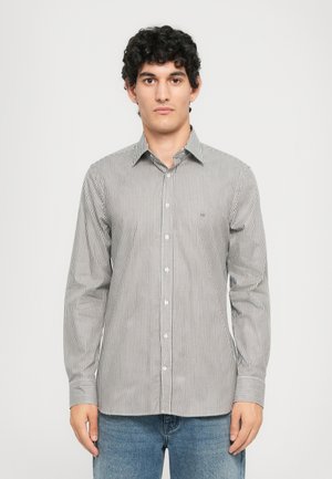 Ung mann med krøllete hår iført en langermet skjorte med vertikale svart-hvite striper og blå jeans mot hvit bakgrunn.
