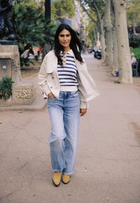 Femme marchant sur un trottoir bordé d'arbres, vêtue d'un pull rayé, d'une veste blanche, de jeans à jambes larges et de bottes jaunes, les mains dans les poches.