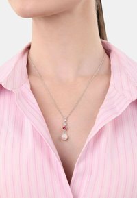 Srebrny naszyjnik z łańcuszkiem, z czerwonym kamieniem i okrągłym zawieszeniem o delikatnym połysku, noszony na różowej bluzce w paski.