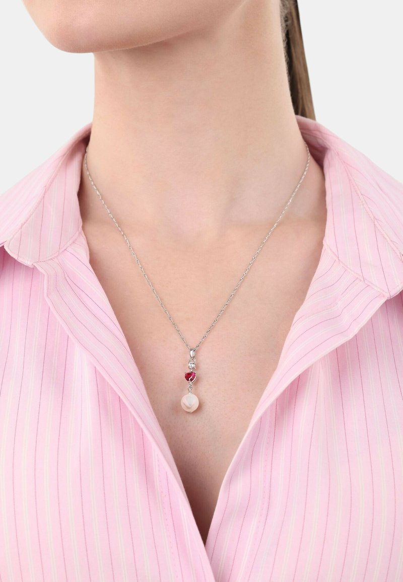 Srebrny naszyjnik z łańcuszkiem, z czerwonym kamieniem i okrągłym zawieszeniem o delikatnym połysku, noszony na różowej bluzce w paski.