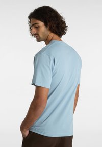 Ljust blå bomullströja med korta ärmar, rund halsringning och en liten svart etikett på vänster ärm. Slät textur, avslappnad passform.