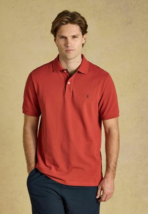 Jonge man met kort bruin haar, gekleed in een rood poloshirt met korte mouwen en een donkere broek, staand tegen een effen beige achtergrond.