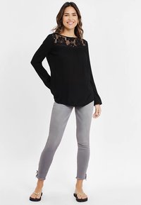 Schwarze langärmelige Bluse mit Spitzendetail am Ausschnitt, kombiniert mit grauen Skinny-Jeans. Das Modell trägt schwarze Sandalen. Schlichter weißer Hintergrund.