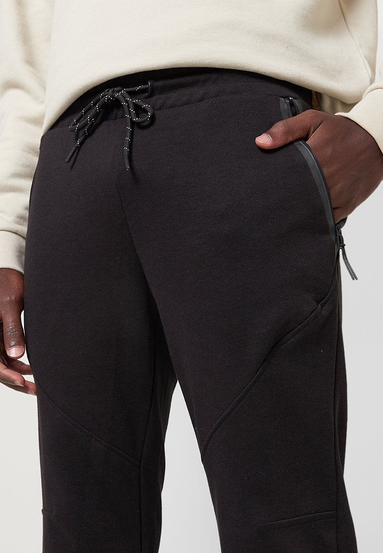 Pantalones de chándal negros con un acabado texturizado, que presentan costuras en zigzag, bolsillos con cremallera y una cinturilla ajustable con cordón.
