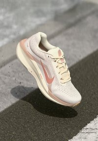 Lichte hardloopschoen in crème met roze accenten. Bovenwerk van mesh, gevoerde zool, afgeronde neus en een opvallend logo aan de zijkant.