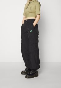 Olivový krátký top s krátkými rukávy a předním zipem, spárovaný s černými cargo kalhotami se zeleným logem Nike. Černé platformové boty.