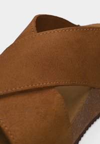 Brun sandal i ruskind med brede krydsremme og en sål i korkplatform mod en neutral baggrund.