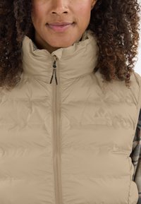 Ljusbeige puffer-väst med hög krage, framzip-stängning och subtil logotyp. Har en vadderad struktur och en mjuk, slät tyg.