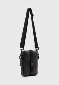 Kompaktní černá kabelka přes rameno s nastavitelným širokým popruhem a předním zapínáním na sponu, zobrazena na jednoduchém světlém pozadí.