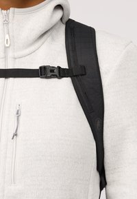 Svart ryggsäcksrem på en grå zip-up-hoodie, med texturerat material och en justerbar spänne på sidan. Närbild som fokuserar på detaljer.